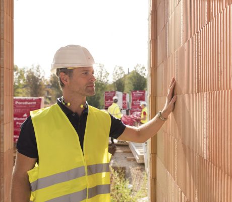 Builder on construction site touching clay block wall, construction workers and clay block pallets in the background